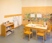 Ein heller Kreativraum mit einem Tisch, vier Stühlen und Malmaterialien. An der Wand hängt eine bemalte Holzplatte. In einer Ecke steht ein Regal mit Bastelutensilien und Kittel. Im Hintergrund ist ein Waschbecken zu sehen.