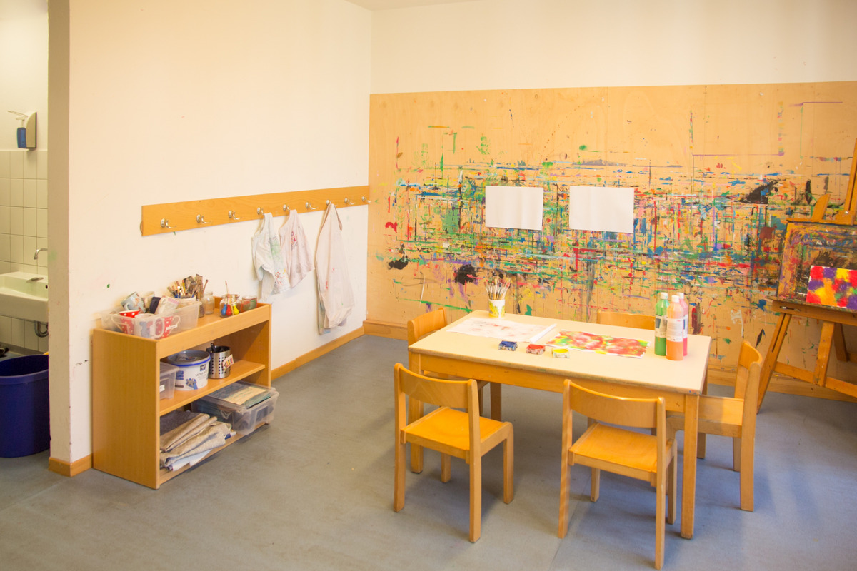 Ein heller Kreativraum mit einem Tisch, vier Stühlen und Malmaterialien. An der Wand hängt eine bemalte Holzplatte. In einer Ecke steht ein Regal mit Bastelutensilien und Kittel. Im Hintergrund ist ein Waschbecken zu sehen.