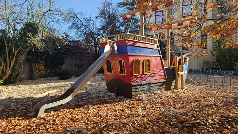 Ein rotes Spielschiff steht auf einem Spielplatz, umgeben von buntem Laub. Es hat eine Rutsche und Fenster. Eine Seilbrücke verbindet das Spielschiff mit dem Boden, der mit herabgefallenen Blättern bedeckt ist. Der Himmel ist blau und es gibt einige kahle Bäume im Hintergrund.