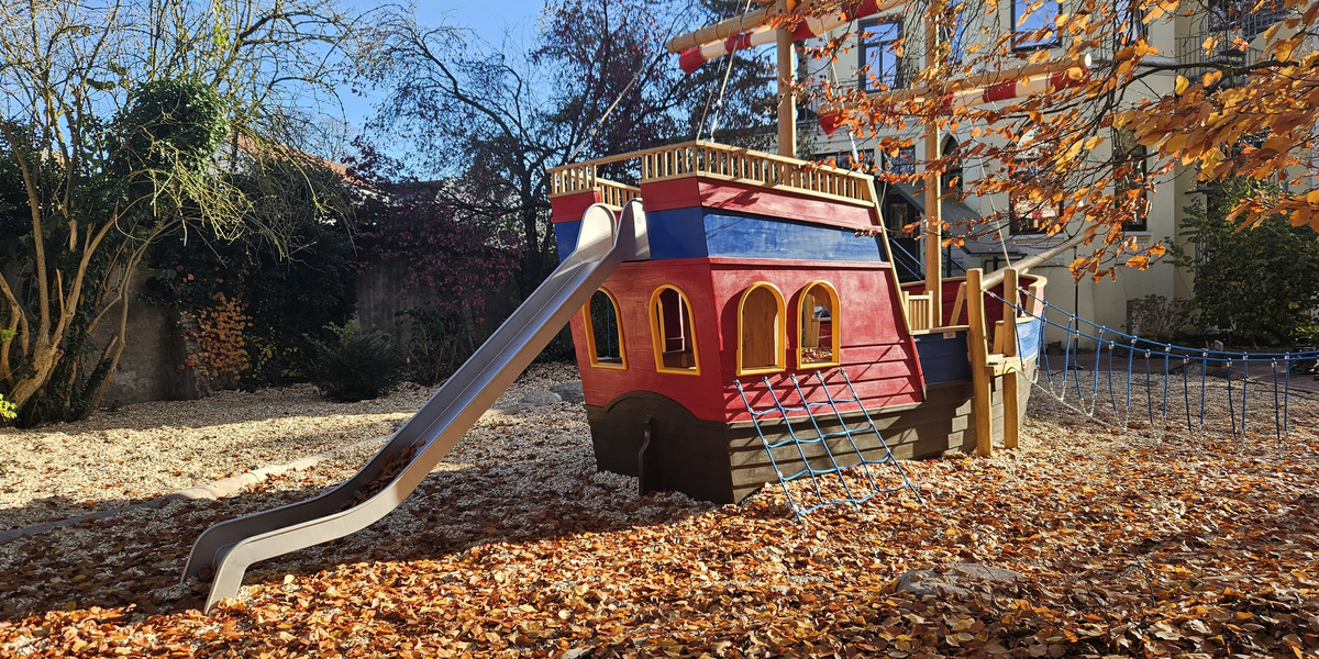 Ein rotes Spielschiff steht auf einem Spielplatz, umgeben von buntem Laub. Es hat eine Rutsche und Fenster. Eine Seilbrücke verbindet das Spielschiff mit dem Boden, der mit herabgefallenen Blättern bedeckt ist. Der Himmel ist blau und es gibt einige kahle Bäume im Hintergrund.