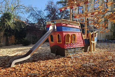 Ein rotes Spielschiff steht auf einem Spielplatz, umgeben von buntem Laub. Es hat eine Rutsche und Fenster. Eine Seilbrücke verbindet das Spielschiff mit dem Boden, der mit herabgefallenen Blättern bedeckt ist. Der Himmel ist blau und es gibt einige kahle Bäume im Hintergrund.
