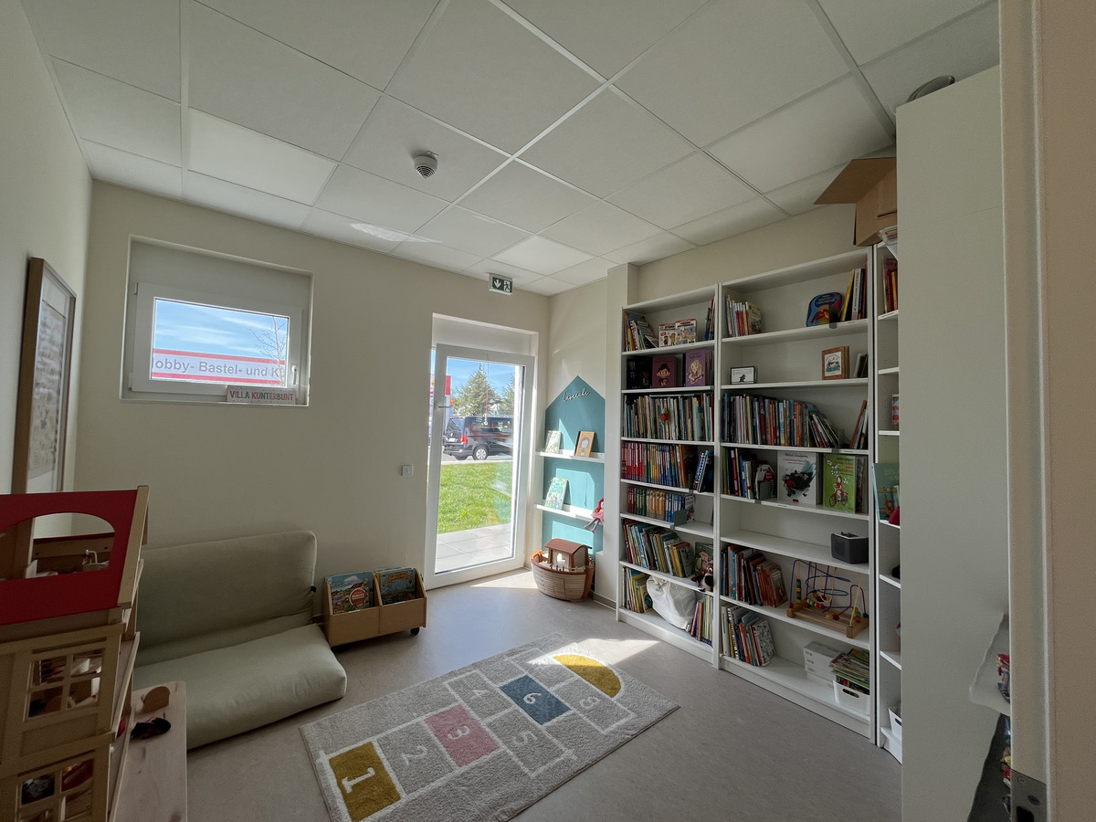 Ein heller Raum mit einem Sofa und einem Teppich. An der Wand steht ein hohes Bücherregal, das mit Büchern gefüllt ist. Ein kleines Spielzeugregal und ein Spielzeugkasten sind sichtbar. Große Fenster lassen Licht herein und bieten einen Blick nach draußen auf einen grünen Bereich.