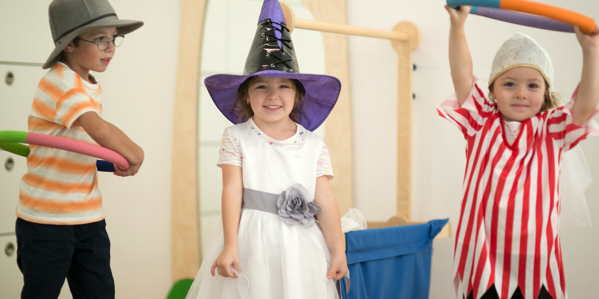 Drei Kinder verkleidet spielen miteinander. Ein Mädchen trägt ein weißes Kleid mit einem lila Zauberhut, während ein Junge in einem gestreiften Shirt mit einem bunten Reifen spielt. Ein weiteres Kind trägt ein historisches Piratenkostüm und hebt ebenfalls einen Reifen. Der Hintergrund zeigt spielerische Elemente.