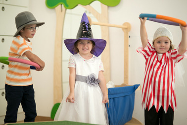 Drei Kinder verkleidet spielen miteinander. Ein Mädchen trägt ein weißes Kleid mit einem lila Zauberhut, während ein Junge in einem gestreiften Shirt mit einem bunten Reifen spielt. Ein weiteres Kind trägt ein historisches Piratenkostüm und hebt ebenfalls einen Reifen. Der Hintergrund zeigt spielerische Elemente.