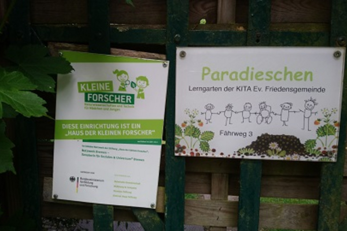 Ein Holzzaun mit zwei Schildern. Links steht „Kleine Forscher“ und informiert über die Einrichtung „Haus der kleinen Forscher“. Rechts befindet sich das Schild „Paradieschen“, das den Lern- und Gartenbereich der Kita Ev. Friedensgemeinde beschreibt.