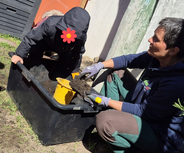 Eine Person mit grauen Handschuhen und blauer Jacke arbeitet mit einem Kind, das in einem schwarzen Behälter sitzt. Beide sind im Freien und beschäftigen sich mit Erde. Im Hintergrund sind verschiedene Gartenutensilien sichtbar.
