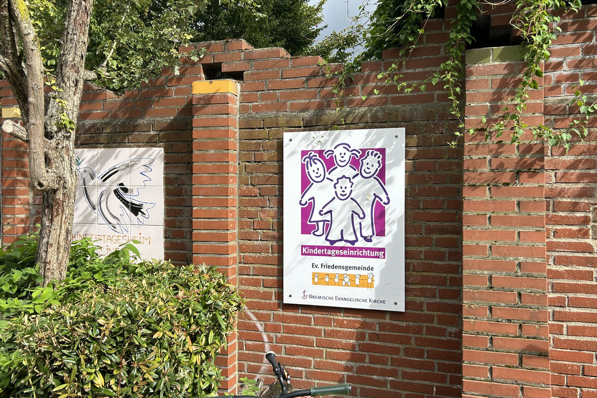 Ein Schild an einer Ziegelwand zeigt vier stilisierte Kinderfiguren in bunten Farben. Der Text darunter lautet "Kindertageseinrichtung Ev. Friedensgemeinde".