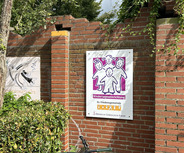 Ein Schild an einer Ziegelwand zeigt vier stilisierte Kinderfiguren in bunten Farben. Der Text darunter lautet "Kindertageseinrichtung Ev. Friedensgemeinde".