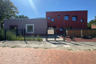 Eine moderne Gebäudeansicht mit einer Kombination aus weißen und roten Wänden. Im Vordergrund ist ein Zaun und ein gepflasterter Weg sichtbar. Einige Grünpflanzen wachsen neben dem Gebäude.
