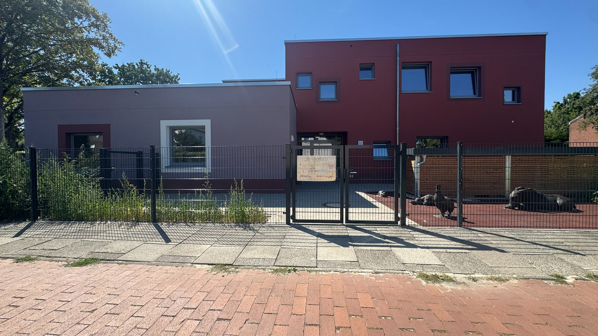 Eine moderne Gebäudeansicht mit einer Kombination aus weißen und roten Wänden. Im Vordergrund ist ein Zaun und ein gepflasterter Weg sichtbar. Einige Grünpflanzen wachsen neben dem Gebäude.