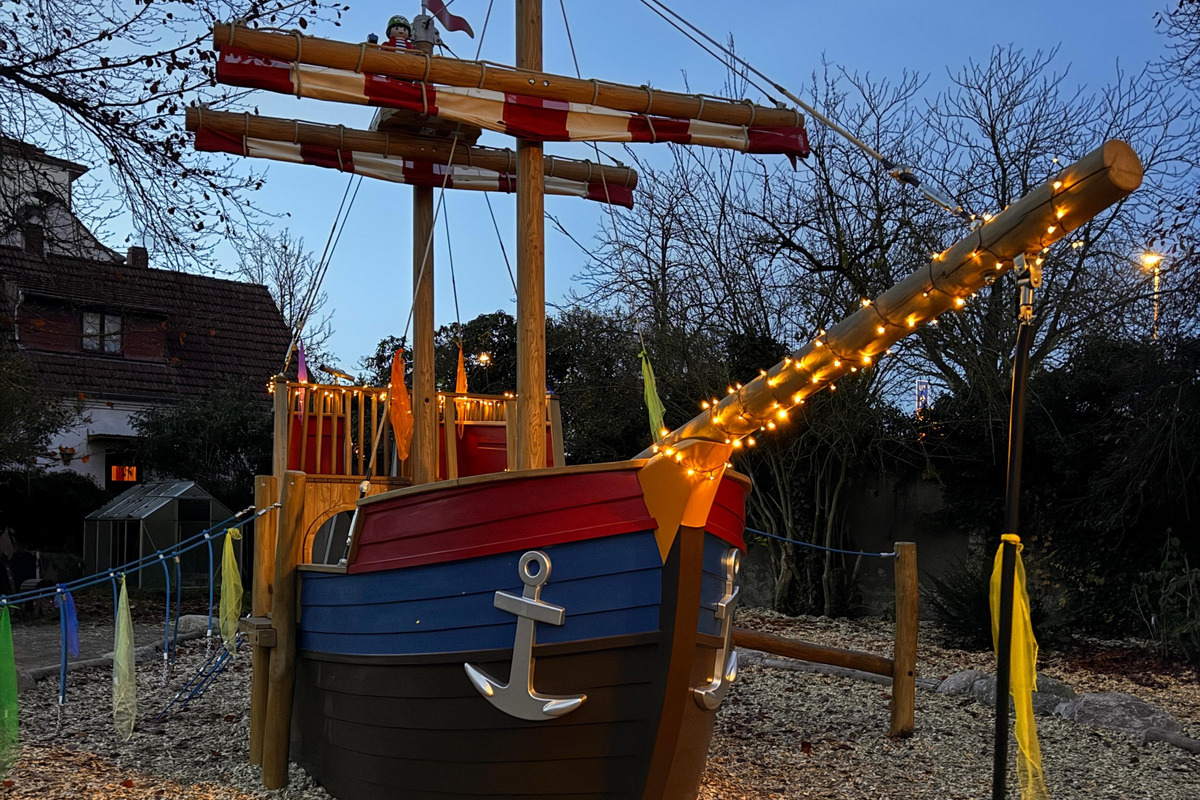 Ein buntes Spielschiff in einem Spielplatzbereich, umgeben von Kies. Es hat ein Piratensymbol auf dem Mast und ist mit Lichtern geschmückt. Der Himmel ist blau und es sind wenige Bäume im Hintergrund zu sehen.