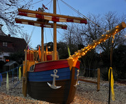 Ein buntes Spielschiff in einem Spielplatzbereich, umgeben von Kies. Es hat ein Piratensymbol auf dem Mast und ist mit Lichtern geschmückt. Der Himmel ist blau und es sind wenige Bäume im Hintergrund zu sehen.