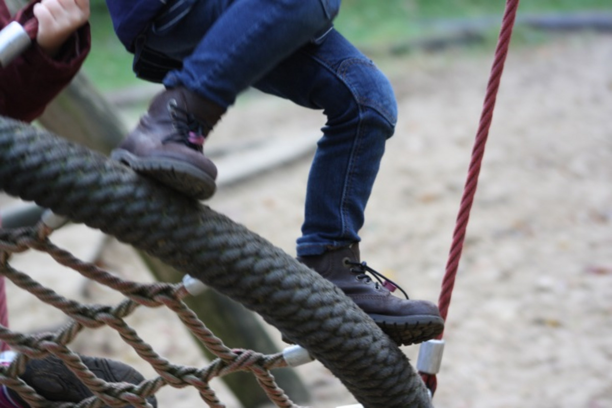 Ein Kind sitzt auf einer schaukelartigen Struktur aus Seilen und Holz auf einem Spielplatz. Es trägt eine Jeans und braune Stiefel, während es mit den Füßen die Kante der Struktur berührt. 