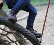 Ein Kind sitzt auf einer schaukelartigen Struktur aus Seilen und Holz auf einem Spielplatz. Es trägt eine Jeans und braune Stiefel, während es mit den Füßen die Kante der Struktur berührt. 