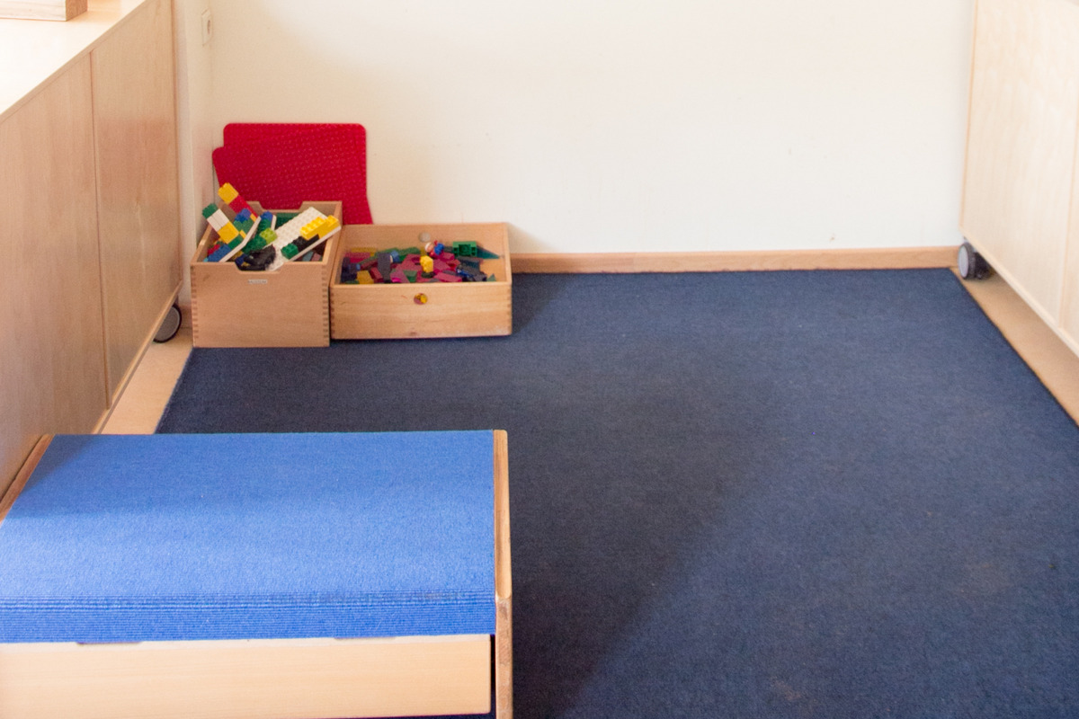 Ein heller Raum mit blauen Teppichboden. An der Wand hängen bunte Papierblumen und ein Informationsbereich mit bunten Karten. Es gibt eine kleine blaue Plattform und zwei Holzkisten mit Spielzeug, darunter Bauklötze. Der Raum wirkt freundlich und einladend für Kinder.