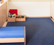 Ein heller Raum mit blauen Teppichboden. An der Wand hängen bunte Papierblumen und ein Informationsbereich mit bunten Karten. Es gibt eine kleine blaue Plattform und zwei Holzkisten mit Spielzeug, darunter Bauklötze. Der Raum wirkt freundlich und einladend für Kinder.