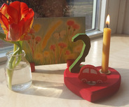 Auf einem Tisch steht ein dekorativer Herzständer mit einer Kerze, einer roten Spielzeugautofigur und der Zahl 2. Daneben befindet sich eine kleine Glasvase mit einer orangefarbenen Tulpe und im Hintergrund ein farbenfrohes Bild von Blumen und Gras.