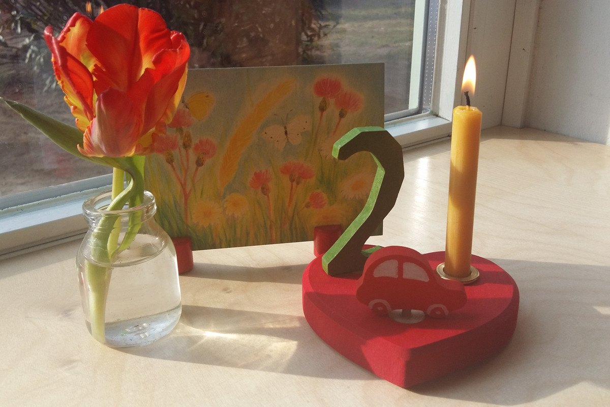Auf einem Tisch steht ein dekorativer Herzständer mit einer Kerze, einer roten Spielzeugautofigur und der Zahl 2. Daneben befindet sich eine kleine Glasvase mit einer orangefarbenen Tulpe und im Hintergrund ein farbenfrohes Bild von Blumen und Gras.