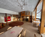 Der Raum ist ein heller, einladender Kindergarten mit Tischen und Stühlen. Im Hintergrund steht ein rotes Pinnbrett mit bunten Bildern. In einer Ecke befindet sich ein erhöhter Spielbereich mit einer Treppe. Der Fußboden ist mit einem runden Teppich gestaltet.