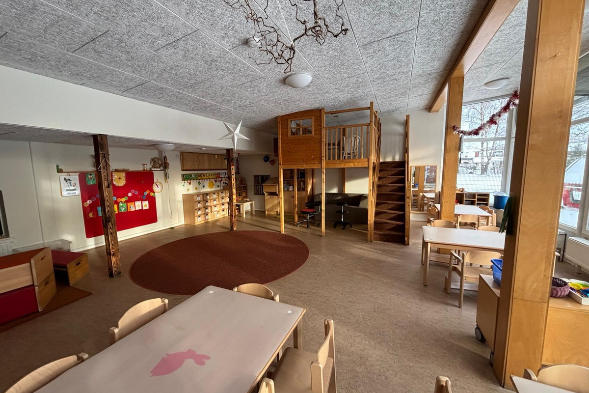 Der Raum ist ein heller, einladender Kindergarten mit Tischen und Stühlen. Im Hintergrund steht ein rotes Pinnbrett mit bunten Bildern. In einer Ecke befindet sich ein erhöhter Spielbereich mit einer Treppe. Der Fußboden ist mit einem runden Teppich gestaltet.