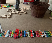 Eine Vielzahl von Spielzeugautos ist in einer Reihe auf einem Teppich angeordnet. Im Hintergrund sind bunte Bauklötze und ein Korb sichtbar. 