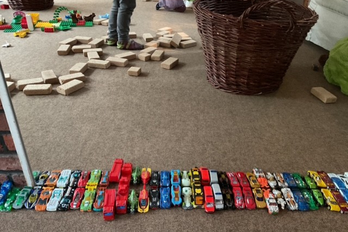 Eine Vielzahl von Spielzeugautos ist in einer Reihe auf einem Teppich angeordnet. Im Hintergrund sind bunte Bauklötze und ein Korb sichtbar. 