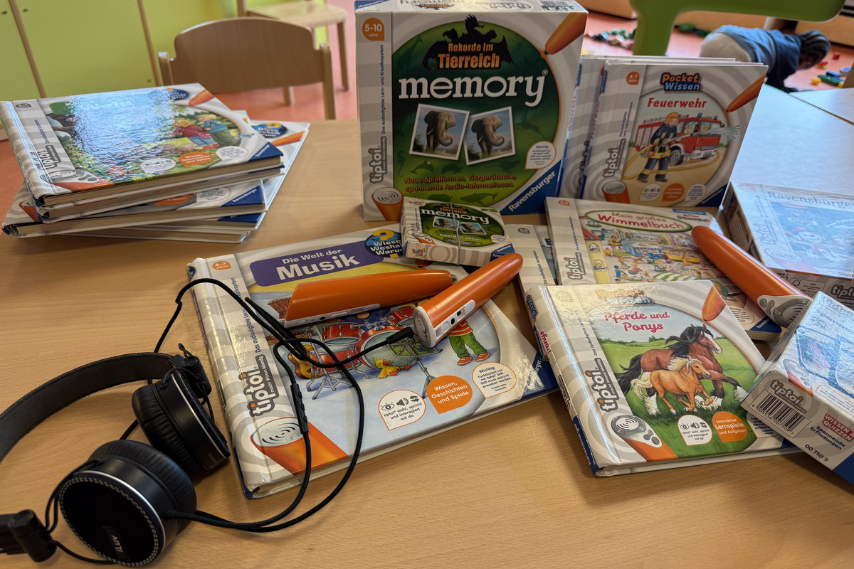 Auf einem Tisch liegen mehrere Kinderbücher und tiptoi-Stifte. Die Bücher umfassen Themen wie Musik, Feuerwehr und Tiere. Neben den Büchern befindet sich ein Paar Kopfhörer, die für die Interaktivität mit den tiptoi-Büchern verwendet werden.