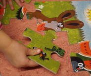 Ein Kind hält ein Puzzlestück mit bunten Tieren und Pflanzenmotiven. Es versucht, das Puzzlestück in ein größeres Puzzle einzufügen. 