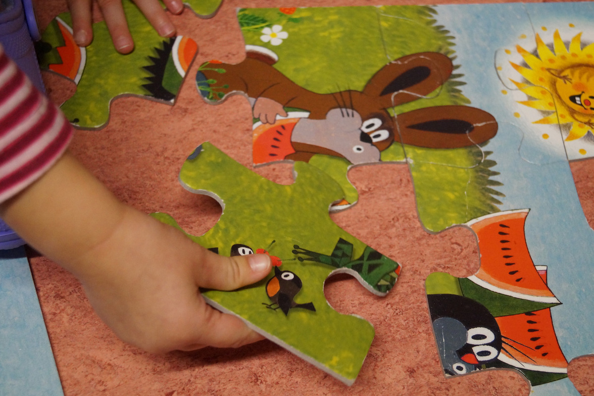Ein Kind hält ein Puzzlestück mit bunten Tieren und Pflanzenmotiven. Es versucht, das Puzzlestück in ein größeres Puzzle einzufügen. 