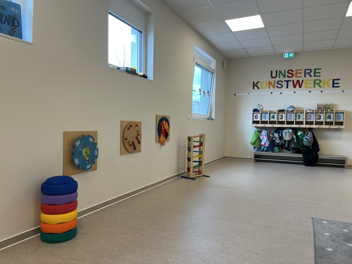 Ein heller Raum mit bunten Wandspielzeugen und einer Garderobe, an der Jacken hängen. An der Wand steht der Schriftzug „UNSERE KUNSTWERKE“. Auf dem Boden stapeln sich große, bunte Sitzkissen in verschiedenen Farben.