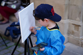 Ein Junge in einem blauen Maleranzug hält einen Bleistift in der Hand und zeichnet auf eine leere Leinwand, die auf einem Ständer steht. In der anderen Hand hält er ein Smartphone. Im Hintergrund sind verschwommene Personen und eine steinerne Wand zu sehen.
