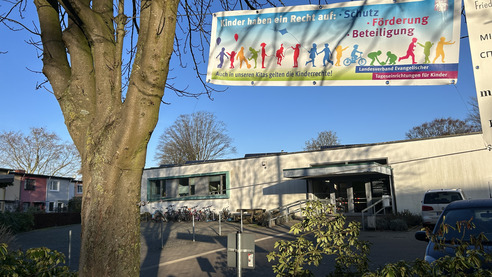 Ein Banner hängt vor einer Einrichtung und betont, dass Kinder ein Recht auf Schutz, Förderung und Beteiligung haben. Darunter steht, dass die Kinderrechte auch in den Kitas gelten. Im Hintergrund ist ein modernes Gebäude mit einem Fahrradabstellplatz zu sehen.