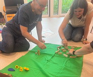 Drei Personen sitzen auf einem grünen Tuch und interagieren mit verschiedenen Spielmaterialien. Es gibt Holzblöcke und einige künstliche Blumen. Im Hintergrund sind weitere Objekte zu sehen.
