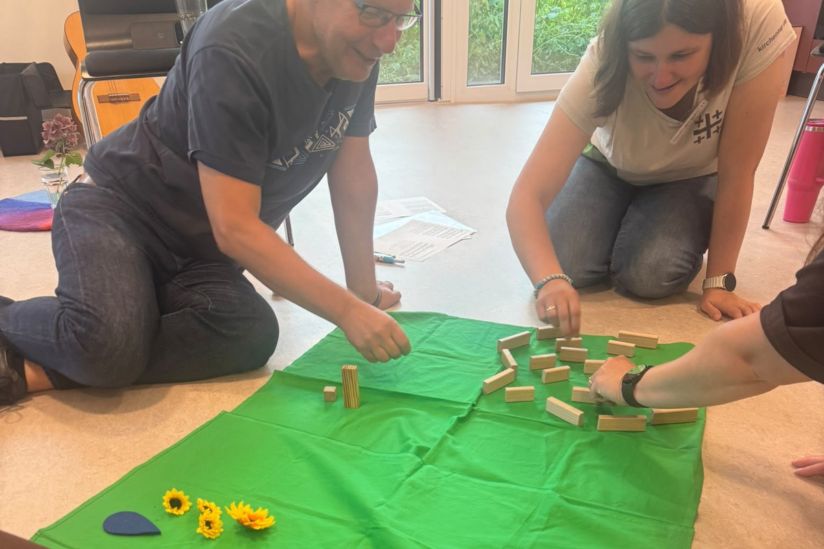 Drei Personen sitzen auf einem grünen Tuch und interagieren mit verschiedenen Spielmaterialien. Es gibt Holzblöcke und einige künstliche Blumen. Im Hintergrund sind weitere Objekte zu sehen.