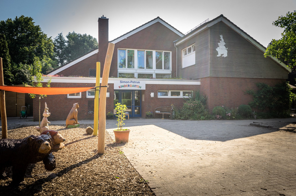 Das Bild zeigt den Eingang des Simon-Petrus Kindergartens. Es ist ein zweistöckiges, ziegelrotes Gebäude mit großen Fenstern. Vor dem Gebäude stehen farbenfrohe Holzskulpturen von Tieren, während der Eingang von einer blühenden Umgebung umgeben ist.