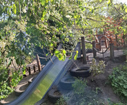 Eine Rutsche aus Metall mit einem sanften Gefälle, umgeben von grünem Gras und alten Reifen. Im Hintergrund ist ein Spielplatz mit Holzkonstruktionen sichtbar. Bäume und Sträucher mit Blüten und Blättern rahmen die Szene ein, was eine naturnahe Atmosphäre schafft.