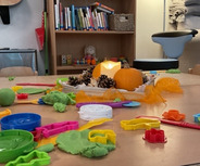 Auf einem Tisch liegen farbige Spielzeuge aus Knete, unter anderem Formen und Kellen. In der Mitte steht ein Kürbis und eine Kerze. Im Hintergrund sind Regale mit Büchern und Spielzeug zu sehen.