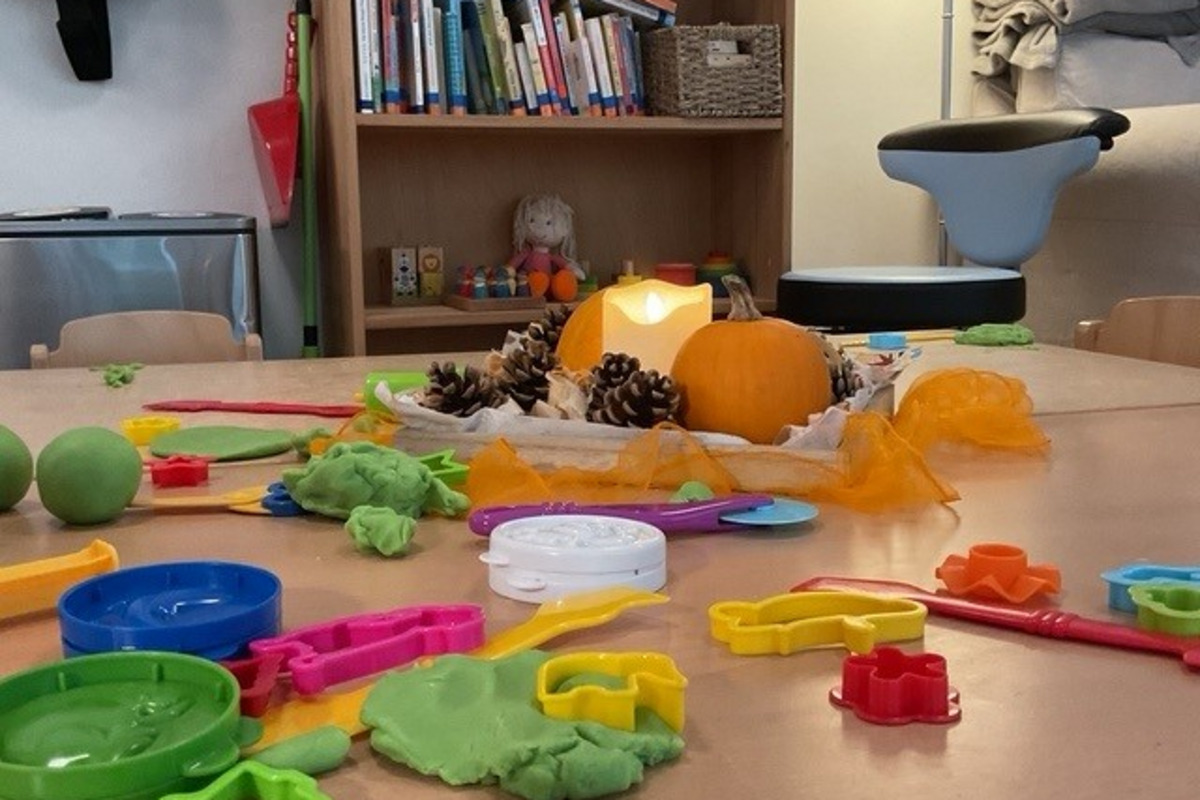 Auf einem Tisch liegen farbige Spielzeuge aus Knete, unter anderem Formen und Kellen. In der Mitte steht ein Kürbis und eine Kerze. Im Hintergrund sind Regale mit Büchern und Spielzeug zu sehen.