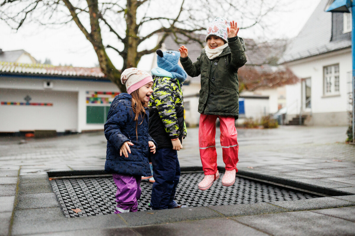 Drei Kinder spielen im Freien. Eines der Kinder springt fröhlich in die Luft, während die anderen lachen. Sie tragen wetterfeste Kleidung und Mützen. Der Boden ist nass, und die Umgebung wirkt freundlich und verspielt.