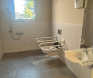 Ein barrierefreies Badezimmer mit einer Dusche, zwei Haltegriffen neben den Toiletten und einem Waschbecken. Es gibt ein Fenster, das Tageslicht hereinlässt, und die Wände sind hell gefliest. Der Boden ist grau.