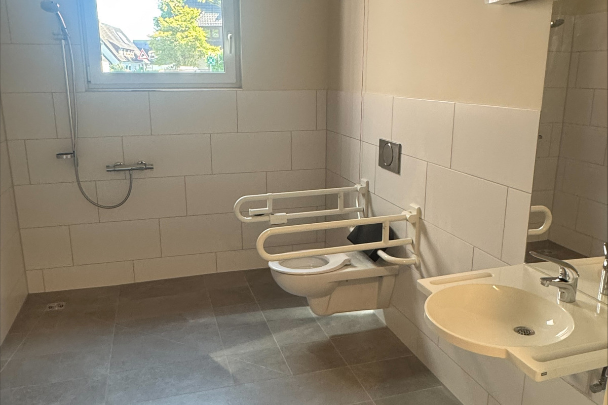 Ein barrierefreies Badezimmer mit einer Dusche, zwei Haltegriffen neben den Toiletten und einem Waschbecken. Es gibt ein Fenster, das Tageslicht hereinlässt, und die Wände sind hell gefliest. Der Boden ist grau.