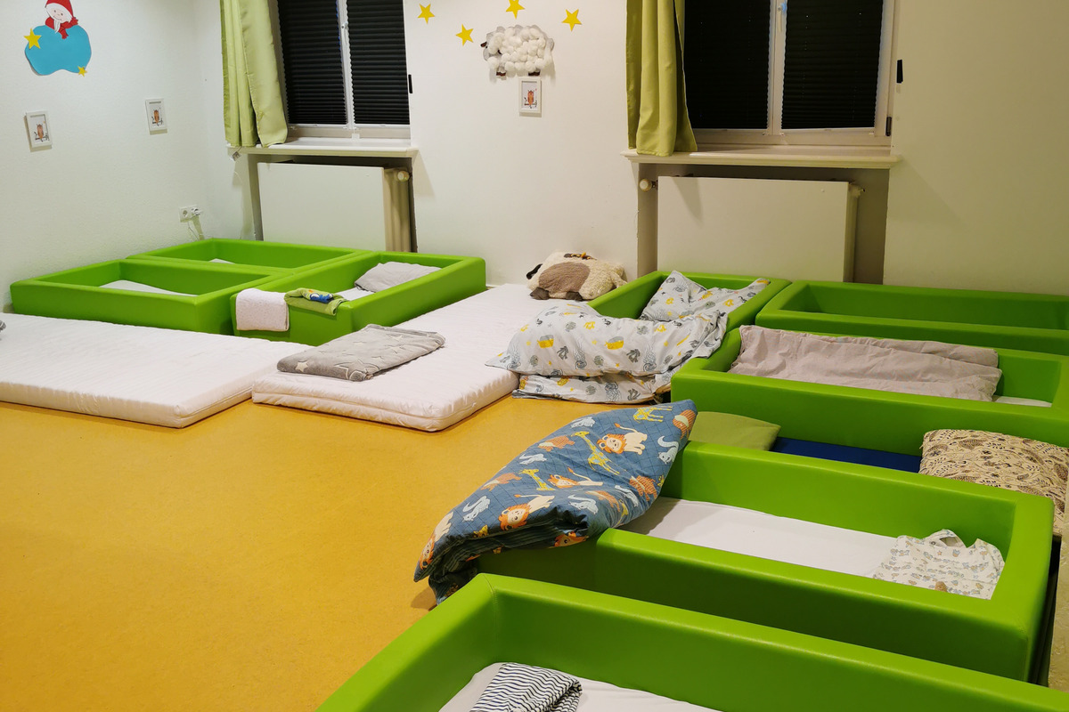 Ein ruhiger Schlafraum mit mehreren grünen Betten und verschiedenen Decken. Die Wände sind hell und mit Sternen sowie einem Bild dekoriert. Vor den Fenstern hängen grüne Vorhänge. Der Boden ist in einem warmen Gelbton gehalten.