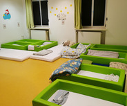 Ein ruhiger Schlafraum mit mehreren grünen Betten und verschiedenen Decken. Die Wände sind hell und mit Sternen sowie einem Bild dekoriert. Vor den Fenstern hängen grüne Vorhänge. Der Boden ist in einem warmen Gelbton gehalten.