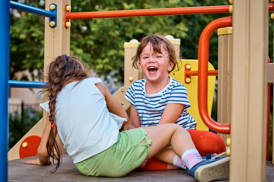 Zwei Kinder spielen auf einem Spielplatz. Ein Mädchen sitzt lachend mit einem gestreiften T-Shirt auf einer roten Fläche, während das andere Mädchen mit einem geflochtenen Zopf daneben sitzt und sich umdreht. Im Hintergrund sind grüne Bäume zu sehen.