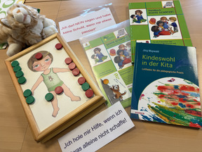 Auf dem Bild sind Materialien zur frühkindlichen Bildung zu sehen. Ein Holzspielbrett mit einer abgebildeten Figur und bunten Chips, zwei Bücher über Kinderschutz und ein Plüschtier liegen auf einem Tisch.