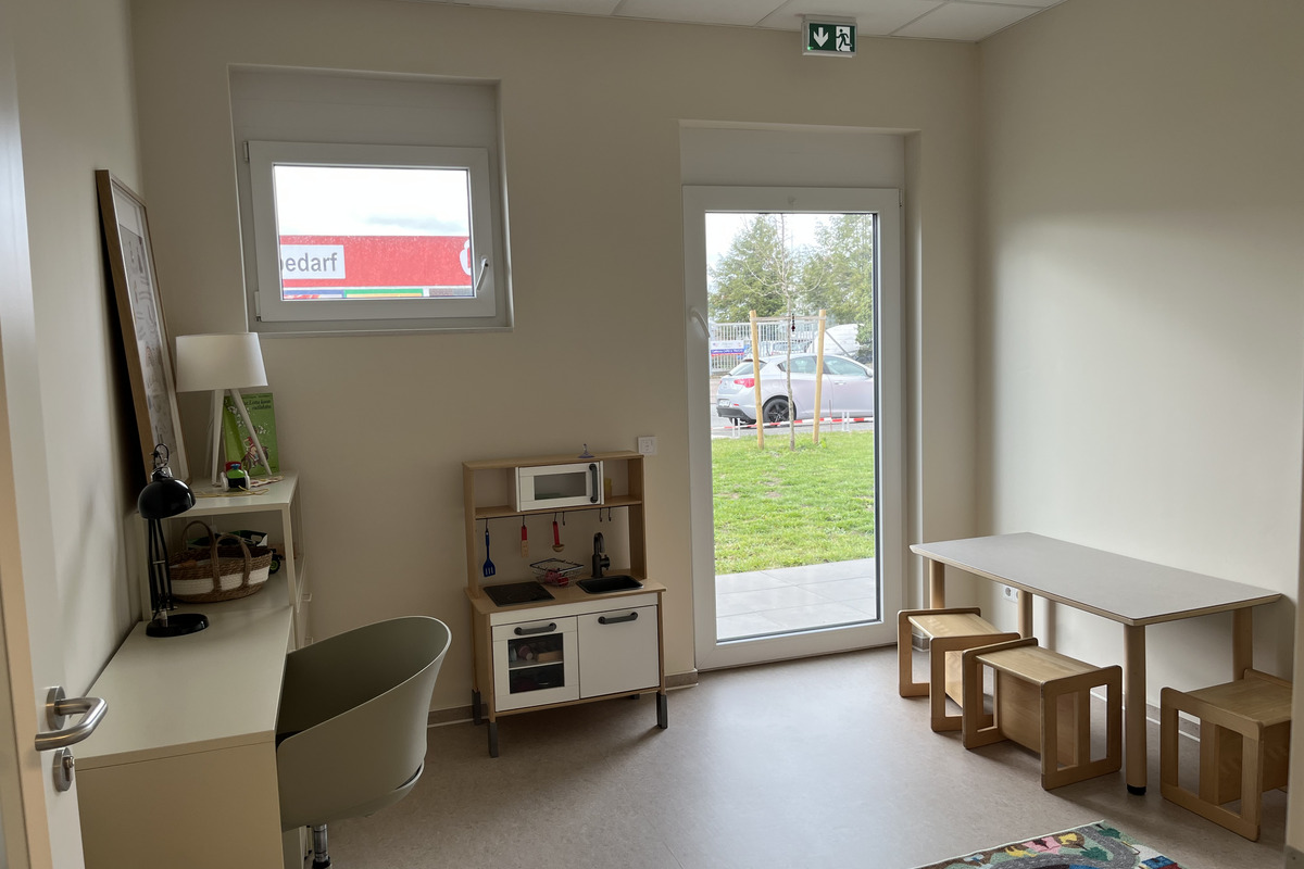 Der Raum ist hell und freundlich gestaltet, mit einer Fensterfront, die auf eine grüne Fläche blickt. Im Raum stehen ein Tisch mit Stühlen, ein kleiner Arbeitsbereich und eine Kinderküche.