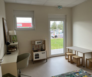 Der Raum ist hell und freundlich gestaltet, mit einer Fensterfront, die auf eine grüne Fläche blickt. Im Raum stehen ein Tisch mit Stühlen, ein kleiner Arbeitsbereich und eine Kinderküche.