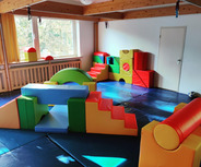 Ein Spielraum mit bunten, weichen Kletterelementen in verschiedenen Formen. Es gibt eine Treppe, Rampen und eine weiche Fläche zum Spielen.