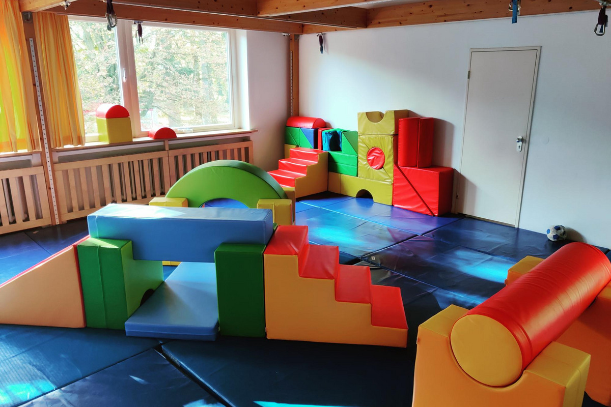 Ein Spielraum mit bunten, weichen Kletterelementen in verschiedenen Formen. Es gibt eine Treppe, Rampen und eine weiche Fläche zum Spielen.