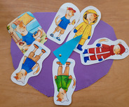 Eine bunte Collage zeigt mehrere Pappfiguren von Katzen in verschiedenen Outfits: eine Katze sitzt und hält Spielzeug, während andere in Regenjacke, Winterkleidung, T-Shirt und Shorts gekleidet sind. Die Figuren sind auf einem lila Hintergrund angeordnet. In der Mitte ist ein Zeiger, der auf eine Katze zeigt.